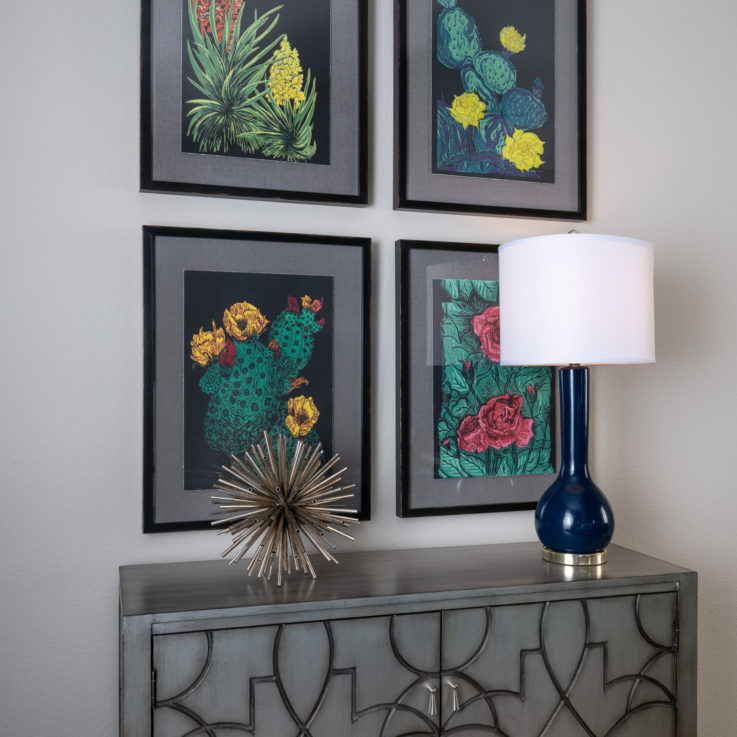 Four flower paintings above a gray cabinet with a blue lamp