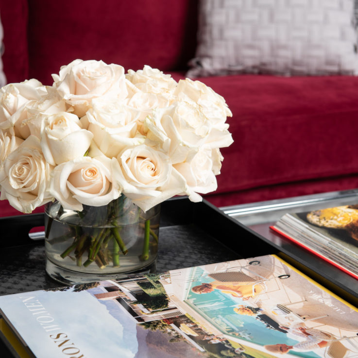 Black tray on a glass coffee table containing interior design books and a glass vase with white roses