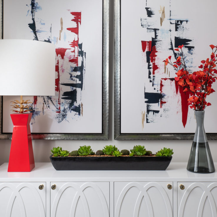 Red lamp and clear vase on white cabinets beneath two abstract paintings