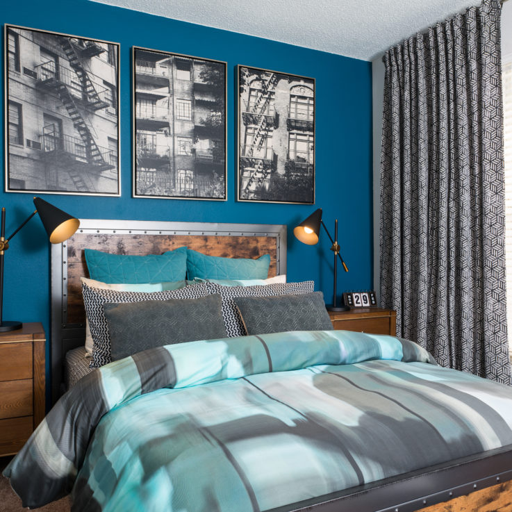 Queen bed with gray, white, and teal covers and two nightstands against a blue wall with three black and white paintings