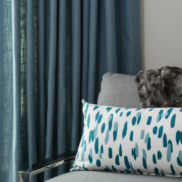 Gray chair with a white and teal pillow in front of a green curtain