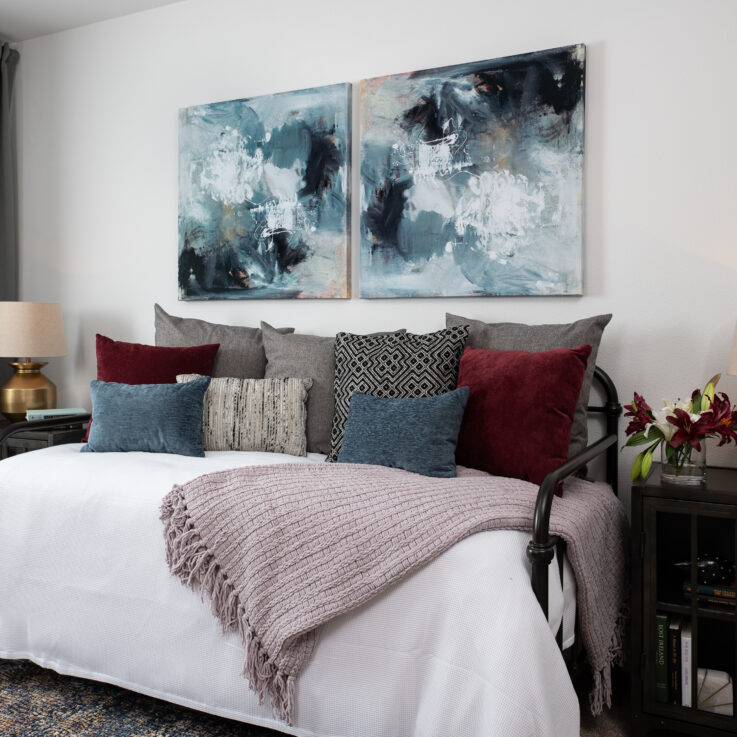 Daybed with blue, purple, and gray pillows below two abstract paintings