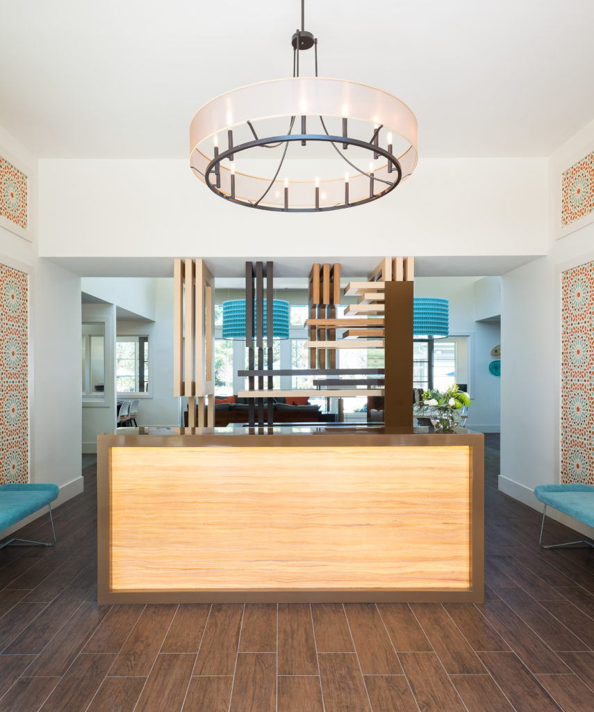 Reception foyer from the leasing lounge featuring various wood finishes, lucite furniture, mixed metals, and bold hues of orange and turquoise in both accents and artwork