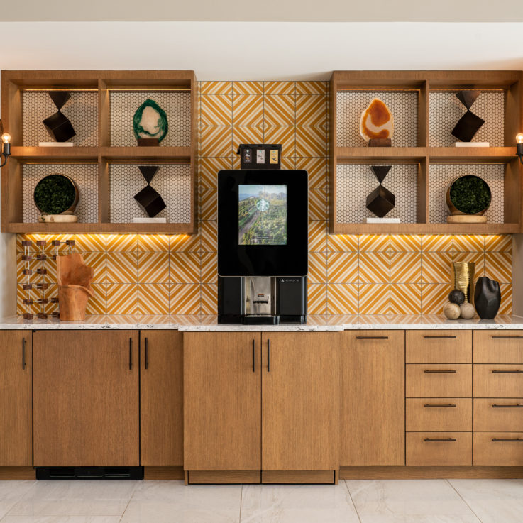 Kitchen area with wooden cabinets, an orange and white tiled wall, and a coffee maker