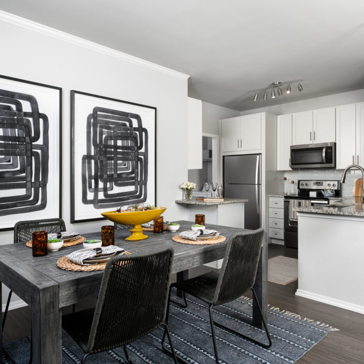 Dining table with four chairs next to a kitchen with white cabinets and stainless steel appliances